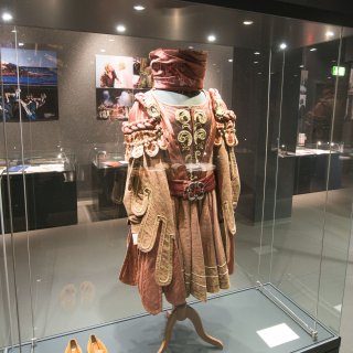Ein rotes Kleid mit goldenen Verzierungen als Ausstellungsstück im Ausstellungsbereich der Abteilung Rheinland in Duisburg. Dazu ein passender Hut und passende Schuhe.