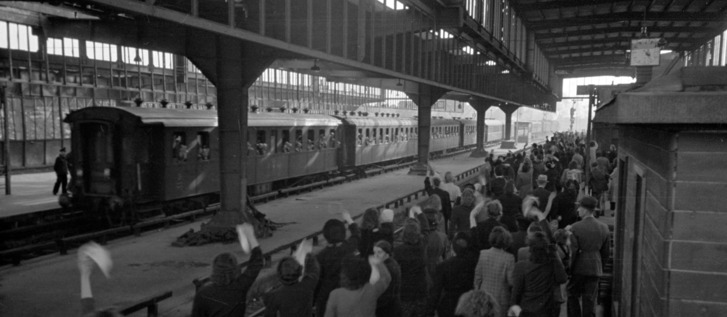 Schwarz Weiß Fotografie eines Bahnsteiges gefüllt mit Menschen. An einem anderen Gleis fährt ein Zug ab.