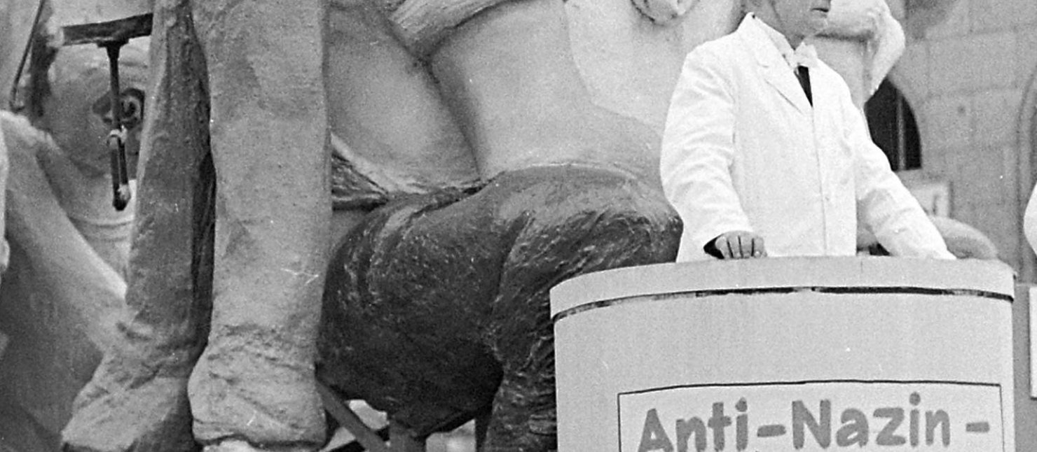 Titelbild der Ausstellung. Zu sehen ist ein Karnevalswagen auf dem "entnazisiert" steht. Ein Patient bekommt eine Spritze.