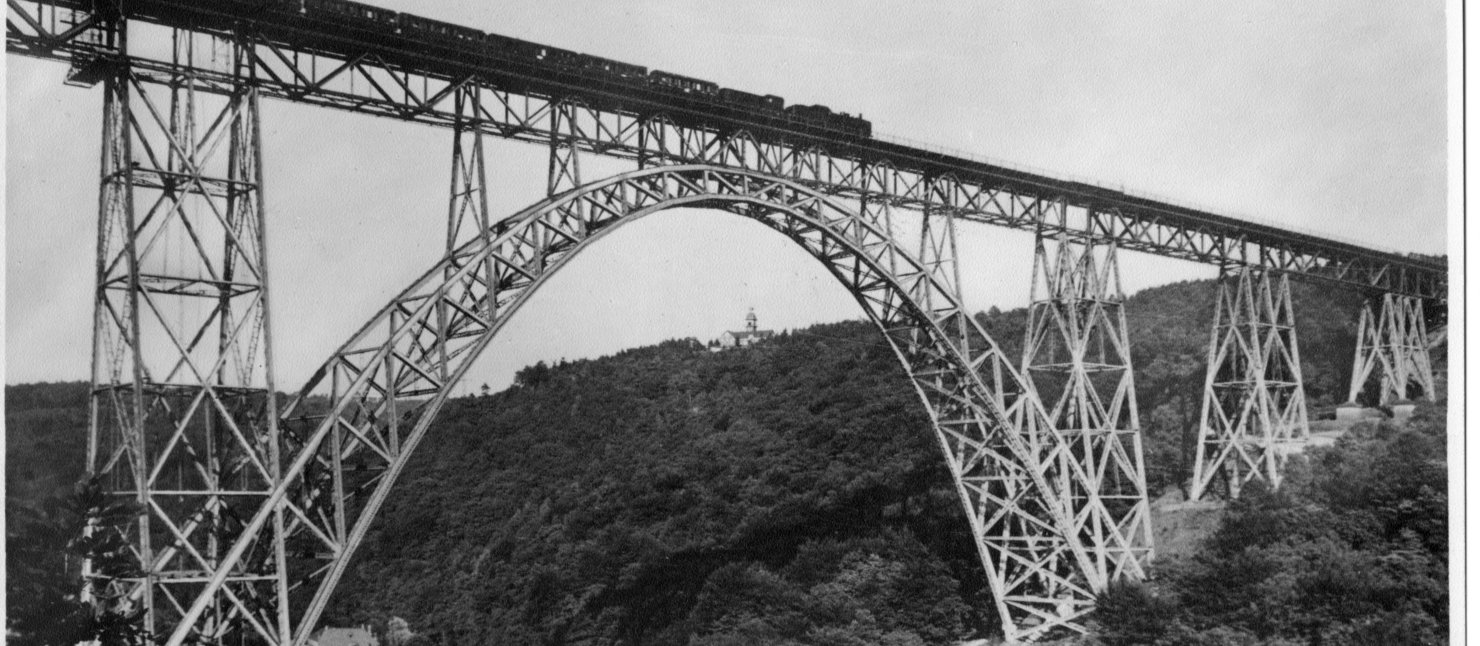 Brücke aus Metall, die über ein Flusstal reicht. Auf der Brücke fährt ein Zug mit Dampflok