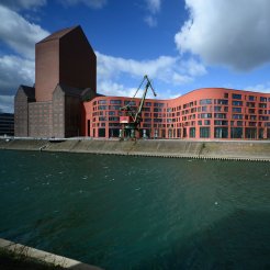 Außenansicht des Dienstgebäudes in Duisburg mit dem Magazinturm und dem wellenförmigen Verwaltungsgebäude, davor zwei Kräne und der Kanal des Innenhafens. 