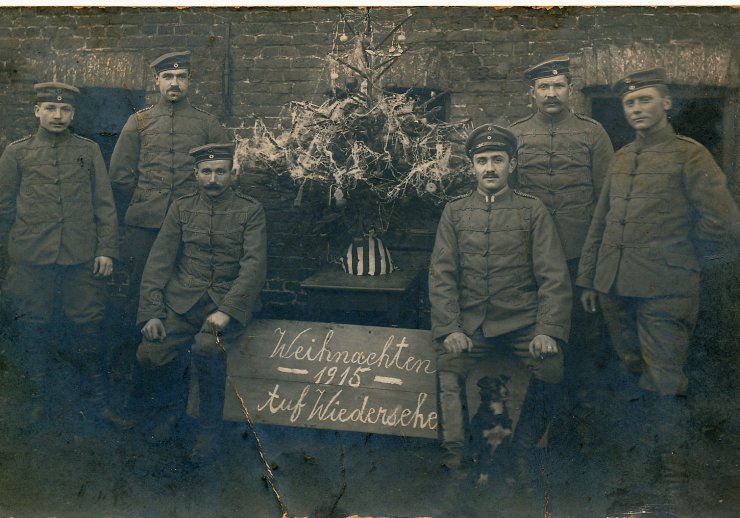 Jeweils drei Männer in Militäruniform stehen rechts und links neben einem kleinen geschmückten Tannenbaum. Unter dem Tannenbaum steht "Weihnachten 1915 Auf Wiedersehen"