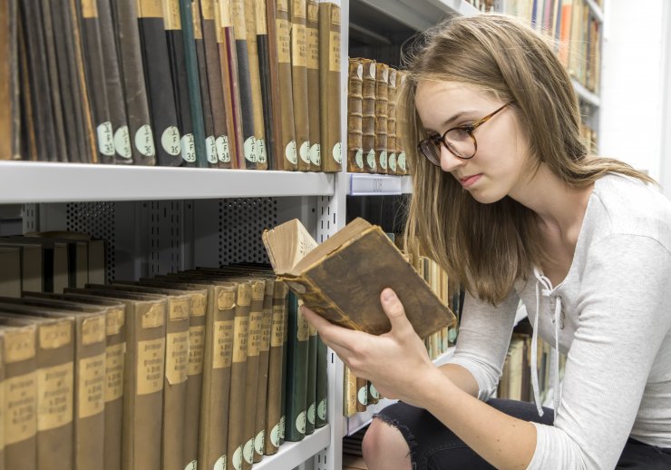Eine junge Frau hockt vor einem Regal voll Bücher. Dabei liest sie in einem besonders alten Buch.