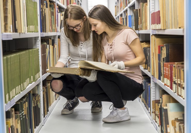 Zwei Praktikantinnen hocken zwischen den mit vielen Archivalien gefüllten Regalen des Landesarchivs. Sie schauen sich zusammen Akten aus den Regalen an.