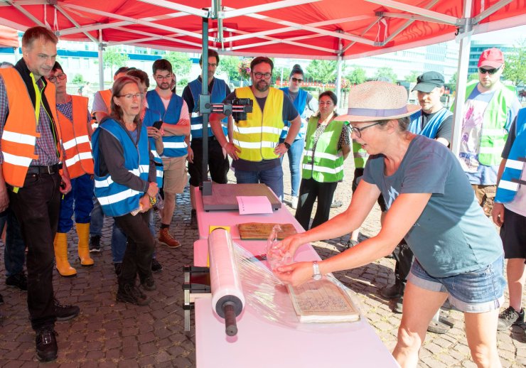 Frau beim einpacken einer Akte in Schutzfolie. Drumherum stehen weitere Personen, die zusehen.