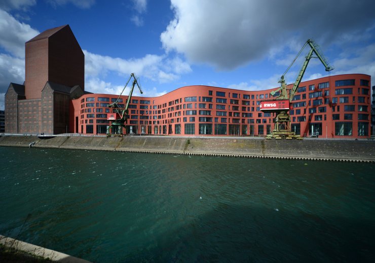 Außenansicht des Dienstgebäudes in Duisburg mit dem Magazinturm und dem wellenförmigen Verwaltungsgebäude, davor zwei Kräne und der Kanal des Innenhafens. 