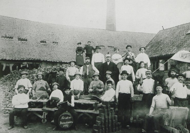 Die Schwarz-weiß-Fotografie aus dem Jahr 1897 zeigt ein Gruppenfoto von zahlreichen Männern in Arbeitskleidung. Im Hintergrund stehen eine Frau und ein Mann auf einem großen Haufen aus Ziegelsteinen etwas höher positioniert. Ein paar der Arbeiter trinken scheinbar alkoholische Getränke. Die Gruppe hat sich auf dem Innenhof der Ziegelei Brunshausen versammelt. Im Vordergrund sind einige Ziegelsteine aufgehäuft, hinter dem Gebäude der Ziegelei ist ein Hochoffen zu erkennen. (LAV OWL D 75 Nr. 2670-6)