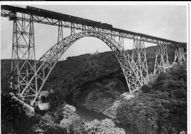 Brücke aus Metall, die über ein Flusstal reicht. Auf der Brücke fährt ein Zug mit Dampflok