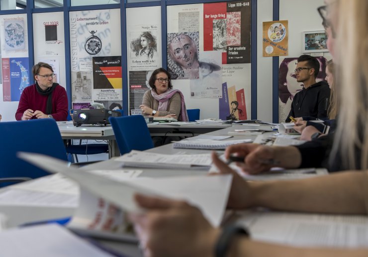 Archivpädagog*innen unterhalten sich mit Besucher*innen in einem Konferenzraum. Dieser ist mit bunten Postern zu geschichtlichen Ausstellungen geschmückt.