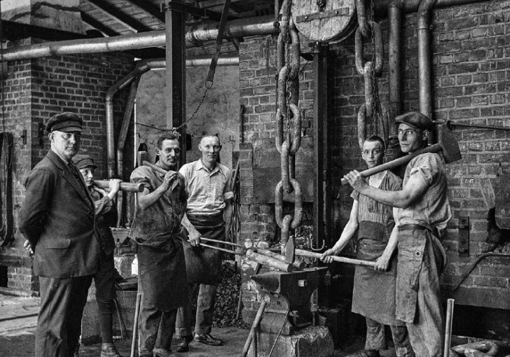 Schwarz-Weiß-Fotografie: 5 Männer stehen mit schwerem Werkzeug und in Arbeitskleidung um einen Amboss.